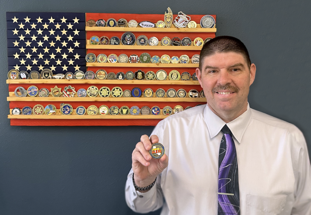 El Jefe Adjunto de la Patrulla de Carreteras de California, Ken Roberts, con una moneda de desafío de la CHP, delante de su colección de monedas de desafío.