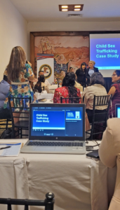 Photo showing the screen of a laptop, people in a classroom setting, and a larger screen at front of a classroom; both screens show the opener for a “Child Sex Trafficking Case Study.”