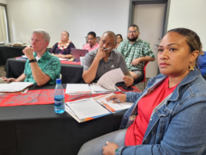 Photo of a woman sitting at a table, with other people behind her, listening in a classroom setting in American Samoa.