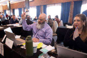 A man at a long conference table ringing a bell in a crowded room