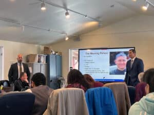 Two men (teachers) near a display screen showing a missing person