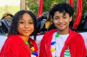 12- and 14-year old sisters wearing fuzzy red hooded jackets with Christmas tree on front and Christmas tree bulb necklaces stand in front of go carts.