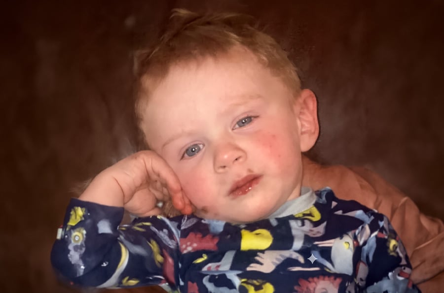 2-year-old boy with blonde hair and blue eyes in dinosaur pajamas resting on furniture