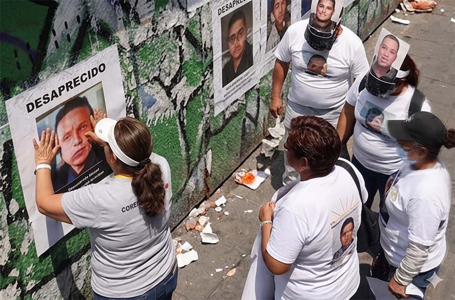 Woman mounting missing person poster on wall in Mexico while others wearing t-shirts and masks with the person's face on surround her and watch.