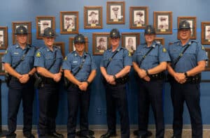 Lieutenant Jim Lowary, Lieutenant Chris Daniels, Lieutenant Morgan Patterson, Captain Jason Crites, Lieutenant Joe Davidson, Lieutenant Casey Utterback in uniform in front of a wall of photos with fallen MSHP fallen officers.