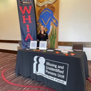 Mary Jo Margeson exhibiting with WHIA banner in back and Missing and Unidentified Persons Unit Washington State Patrol table drape.