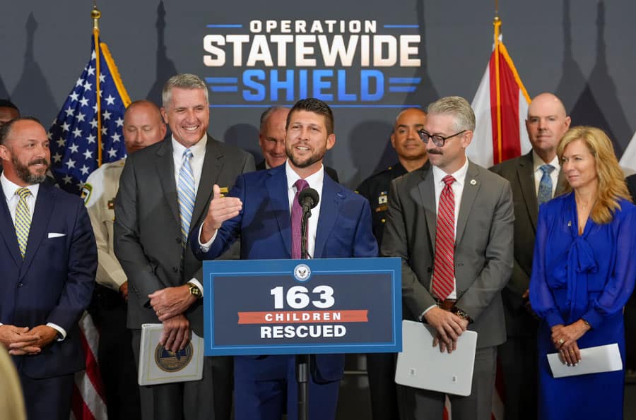 Fiscal General de Florida en la rueda de prensa de la OPERACIÓN STATEWIDE SHIELD con un cartel en el podio que dice '163 NIÑOS RESCATADOS'.