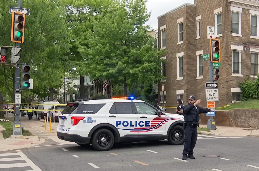 Police officer directing traffic form middle of intersection. Police SUV and crime scene tape block a tree-lined street in front of brownstone brick townhouses.